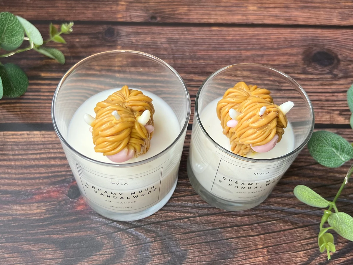 Two Myla Soaps Highland Cow candles in glass jars, each topped with a Highland Cow wax figure, seen from above on a wooden table